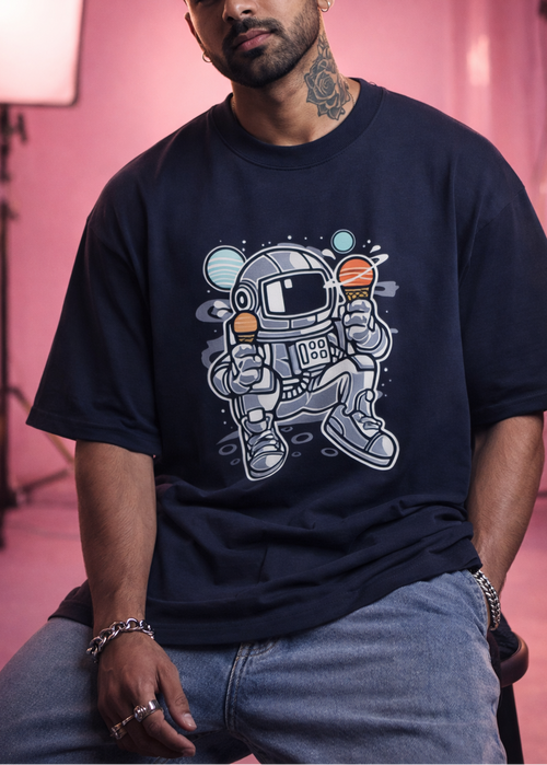Man wearing a navy blue t-shirt with an astronaut graphic holding ice cream cones, sitting against a pink background.