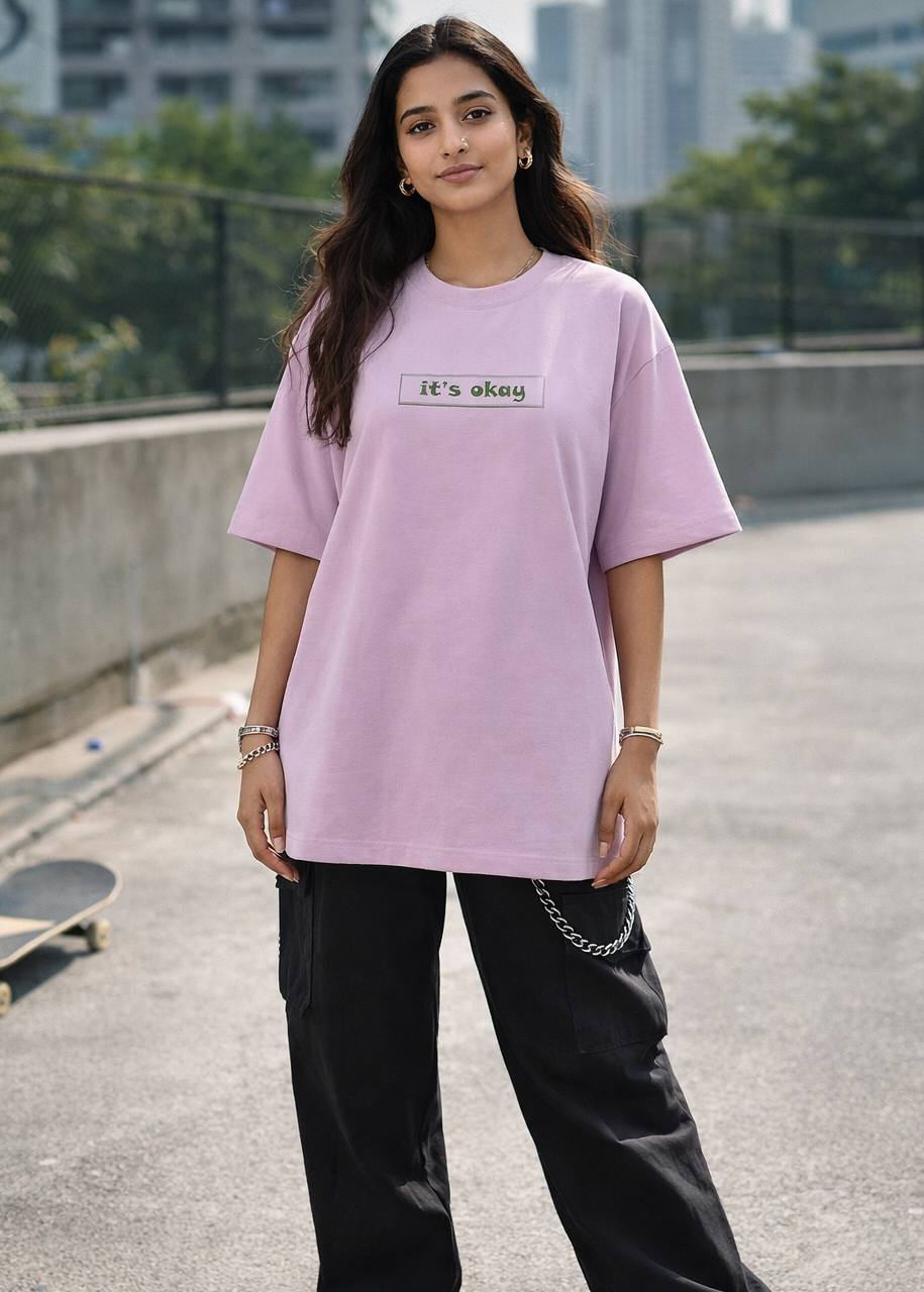 Person wearing a light purple t-shirt with text, standing on a rooftop with a skateboard and cityscape in the background.