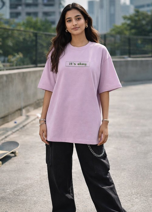 Person wearing a light purple t-shirt with text, standing on a rooftop with a skateboard and cityscape in the background.